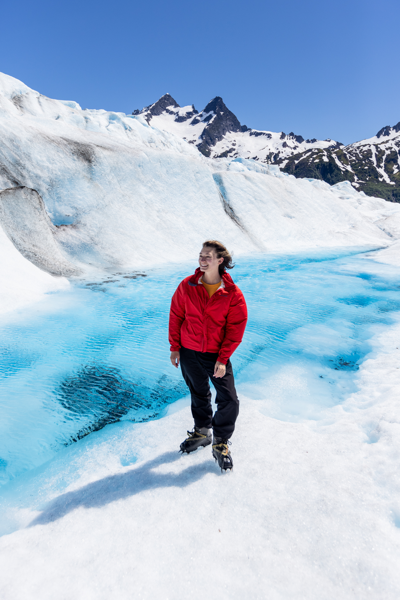 Lindsay, One Girl Wandering, in a red jacket stands in front of a bright blue pond on Mendenhall Glacier