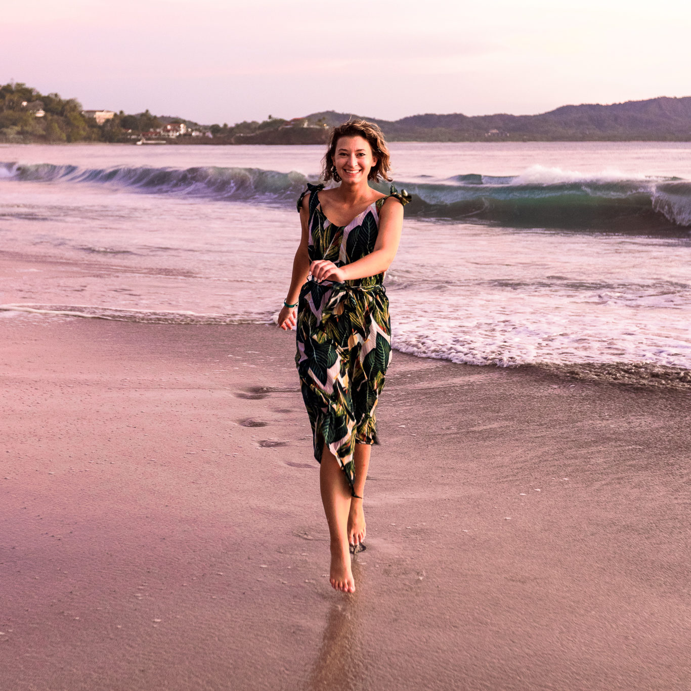 Woman wearing tropical jumpsuit runs towards camera smiling on a beach in Costa Rica. Outfit is part of a Costa Rica vacation lookbook.