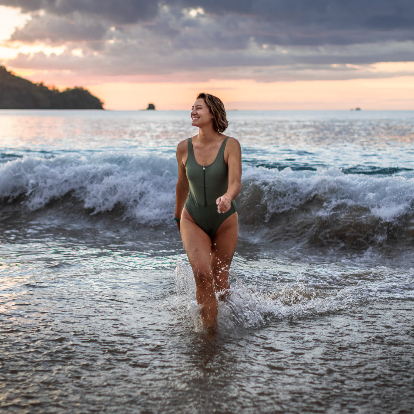 Woman wearing green swimsuit walks through ocean waves at sunset in Costa Rica. Outfit is part of a Costa Rica vacation lookbook.
