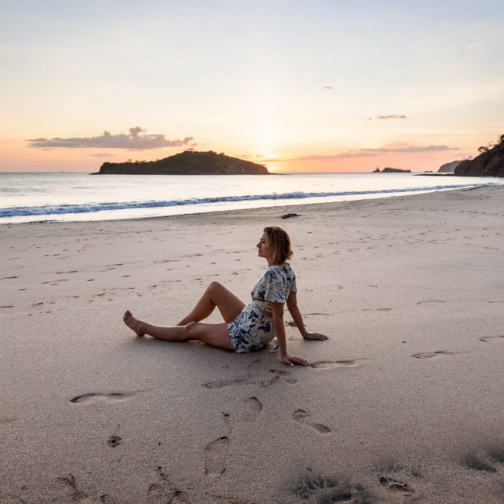 Woman sits on beach in matching two piece outfit looking at the sunset on Costa Rican beach. Outfit is part of a Costa Rica vacation lookbook.
