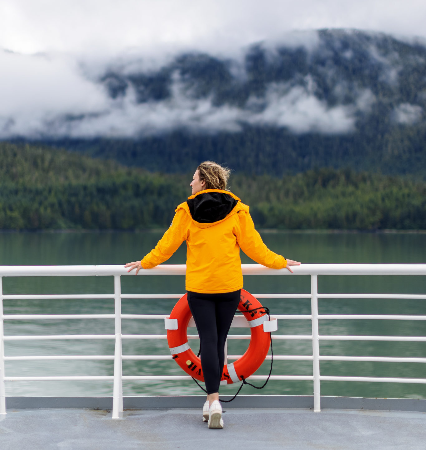 A woman stands on the deck of a ferry in Alaska wearing a bright yellow jacket and black leggings. Pack a good coat when you visit Alaska.