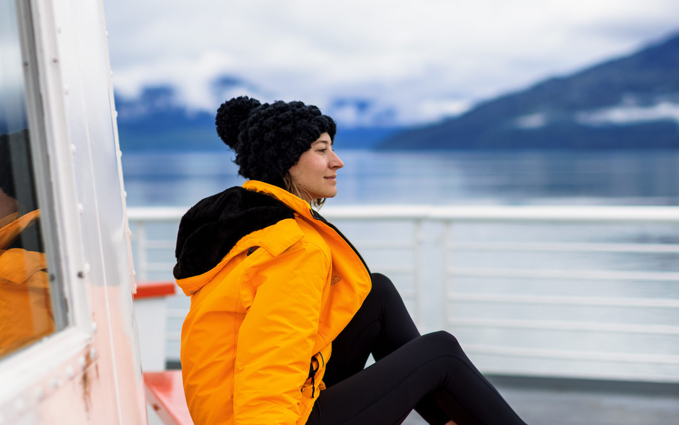 A woman wearing a yellow coat and black leggings sits on an Alaskan ferry